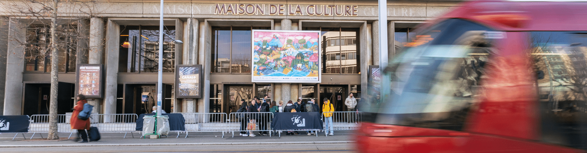 Vue sur la façade avant de la Maison de la culture habillée de l'affiche du festival 2024, un tram s'apprête à passer devant